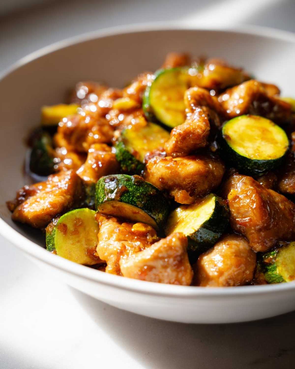 Close-up of glossy, saucy chicken zucchini stir fry served in a white bowl under bright light.