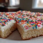 Close-up of thick, soft Christmas sugar cookie bars topped with white frosting and colorful rainbow sprinkles.