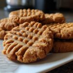 Close-up of several golden brown peanut butter cookies stacked on a white plate, showing classic fork marks.