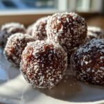 Close-up of several rich, dark bourbon balls rolled in shredded coconut, sitting on a white plate.