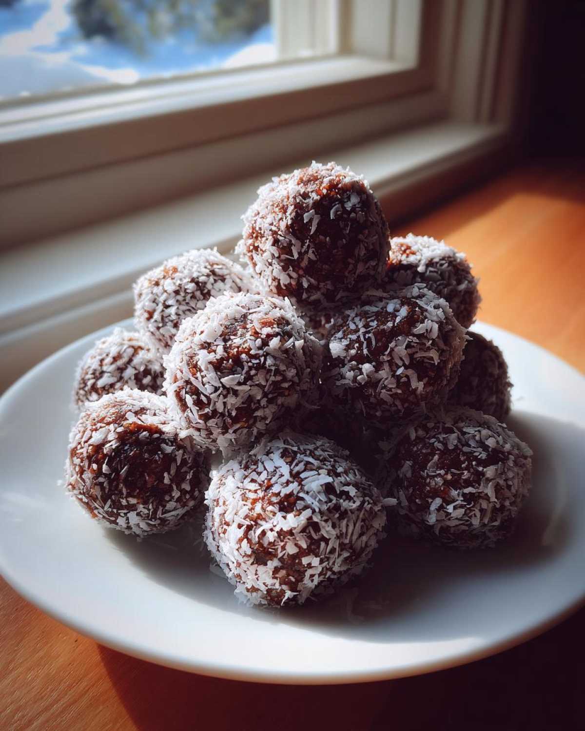 A pile of rich, dark bourbon balls rolled in shredded coconut, sitting on a white plate near a sunlit window.