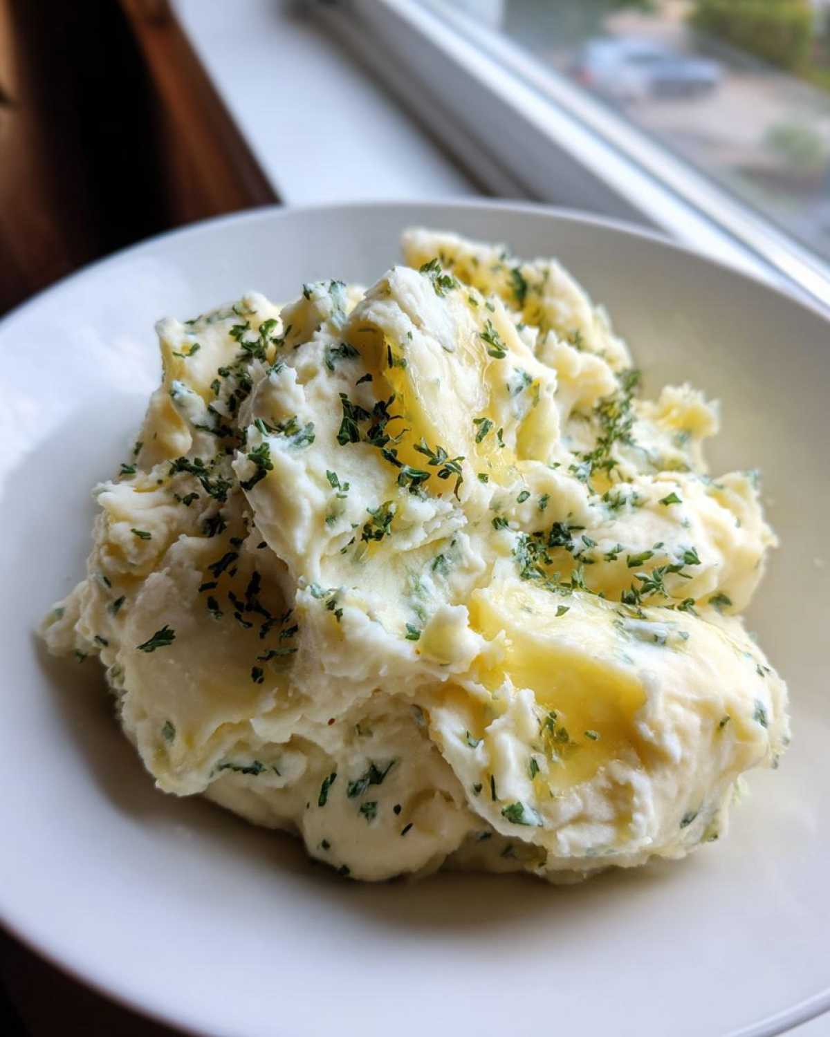 A close-up of creamy Boursin mashed potatoes topped with melted butter and fresh chopped parsley in a white bowl.