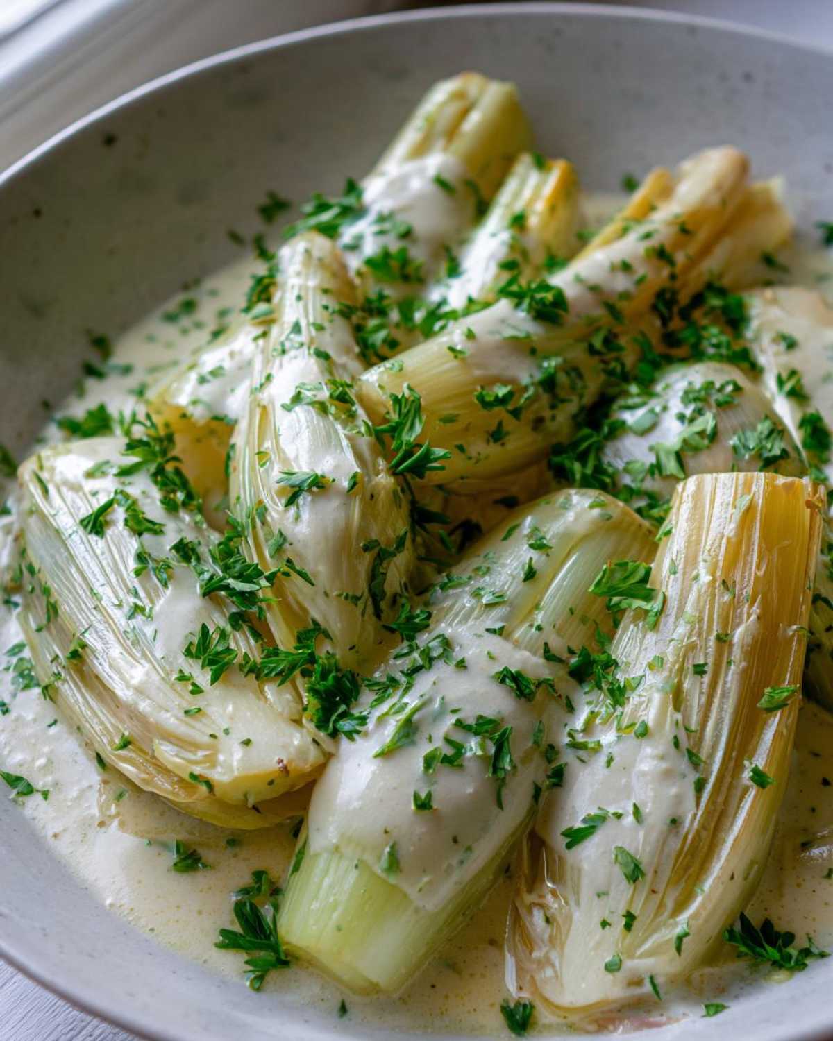 Close-up of tender braised leeks covered in a creamy white sauce and sprinkled generously with fresh chopped parsley.