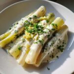 Four tender stalks of braised leeks covered in a creamy white sauce and topped with fresh parsley.