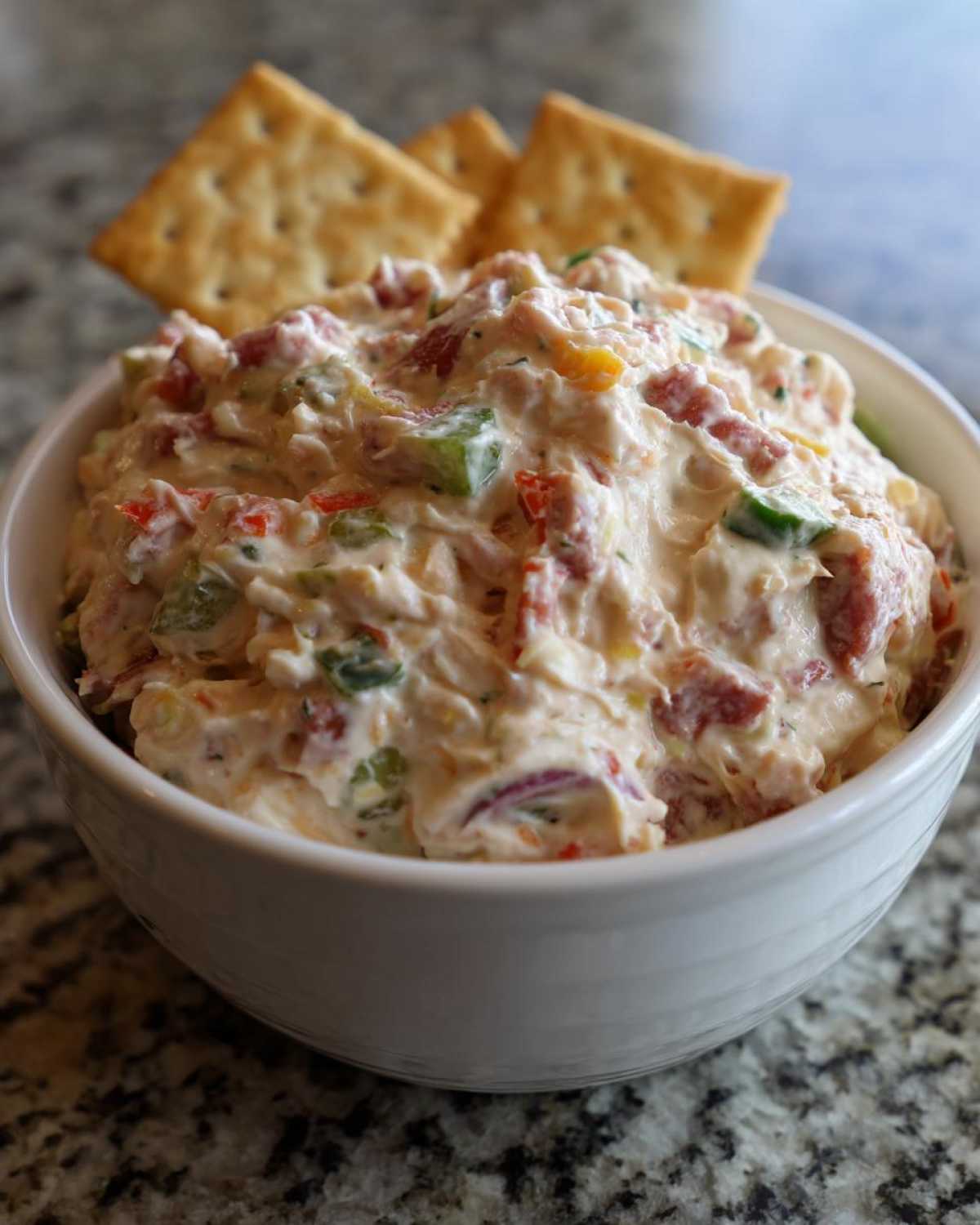 A white bowl filled with creamy hoagie dip mixed with meat and vegetables, served with crackers.