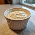 A small white ramekin filled with thick, creamy horseradish sauce speckled with green herbs.