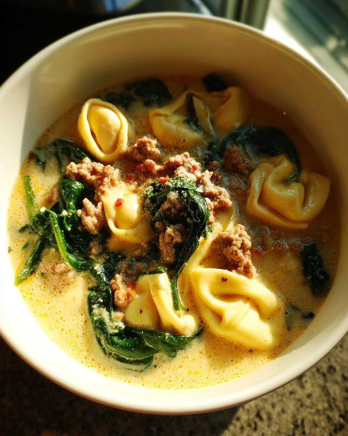 Close-up of a white bowl filled with creamy sausage tortellini soup, featuring tortellini pasta, crumbled sausage, and wilted spinach.