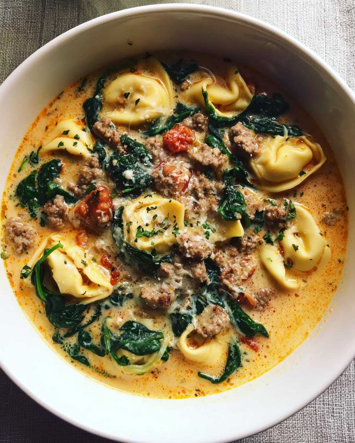 A close-up overhead view of a white bowl filled with creamy sausage tortellini soup, spinach, and ground sausage.