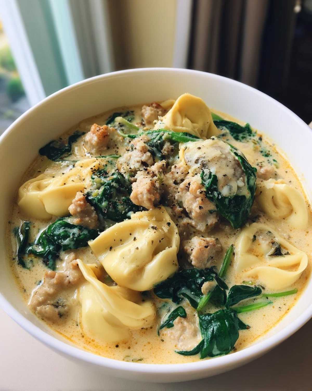 Close-up of a white bowl filled with creamy sausage tortellini soup, featuring pasta, crumbled sausage, and wilted spinach.