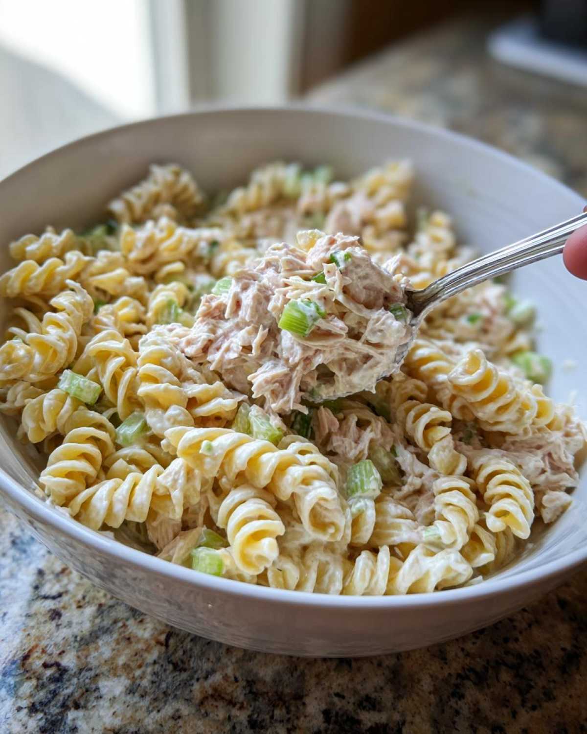A spoonful of creamy tuna pasta salad, featuring rotini pasta and chopped celery, being lifted from a white bowl.