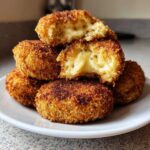 A stack of crispy, golden-brown potato croquettes, one broken open to show a gooey, cheesy interior.