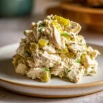 A mound of creamy dill pickle chicken salad featuring chunks of chicken and bright green pickle pieces, served on a light plate.