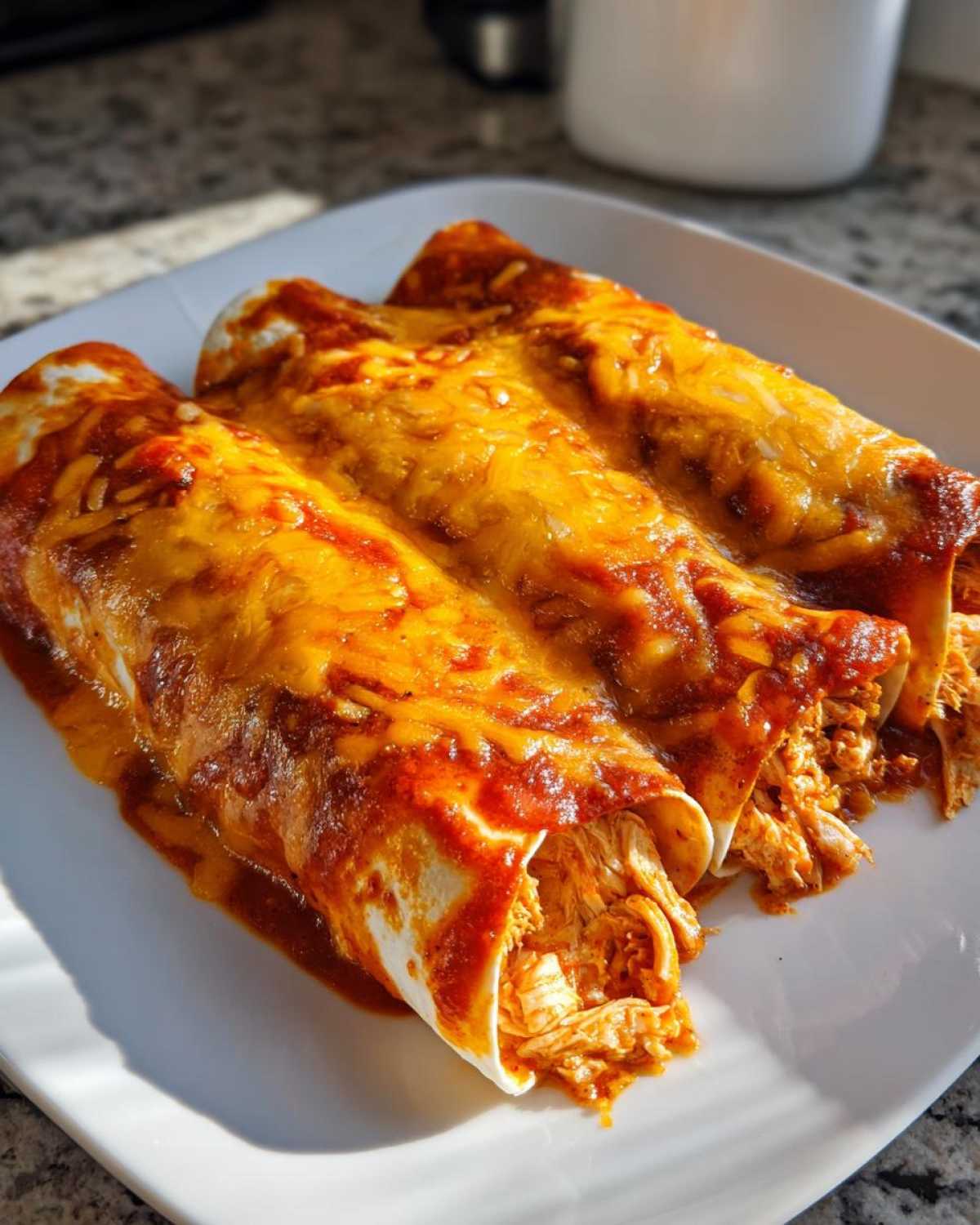 Three rolled tortillas filled with shredded chicken, smothered in red sauce and melted cheddar cheese, representing easy chicken enchiladas.