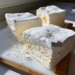 Three fluffy squares of homemade marshmallows dusted heavily with powdered sugar on a white cutting board.