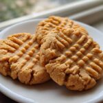 Three classic, fork-pressed Easy Peanut Butter Cookies resting on a white plate near a window.