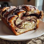 A close-up of a freshly baked chocolate babka loaf, showing rich swirls of chocolate filling and white icing on top.