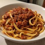 A close-up of fettuccine pasta generously topped with thick, rich bolognese sauce in a white bowl.