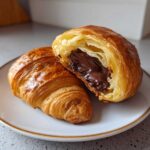 A flaky, golden-brown chocolate croissant cut in half, revealing a rich, melted chocolate filling inside.