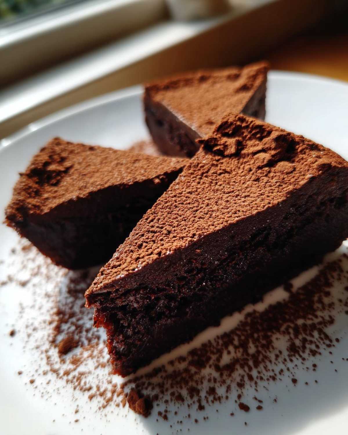 Three triangular slices of dense, dark flourless chocolate torte dusted heavily with cocoa powder on a white plate.