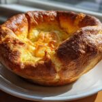 A single, golden brown, perfectly risen yorkshire pudding sitting on a white plate, lit by sunlight.