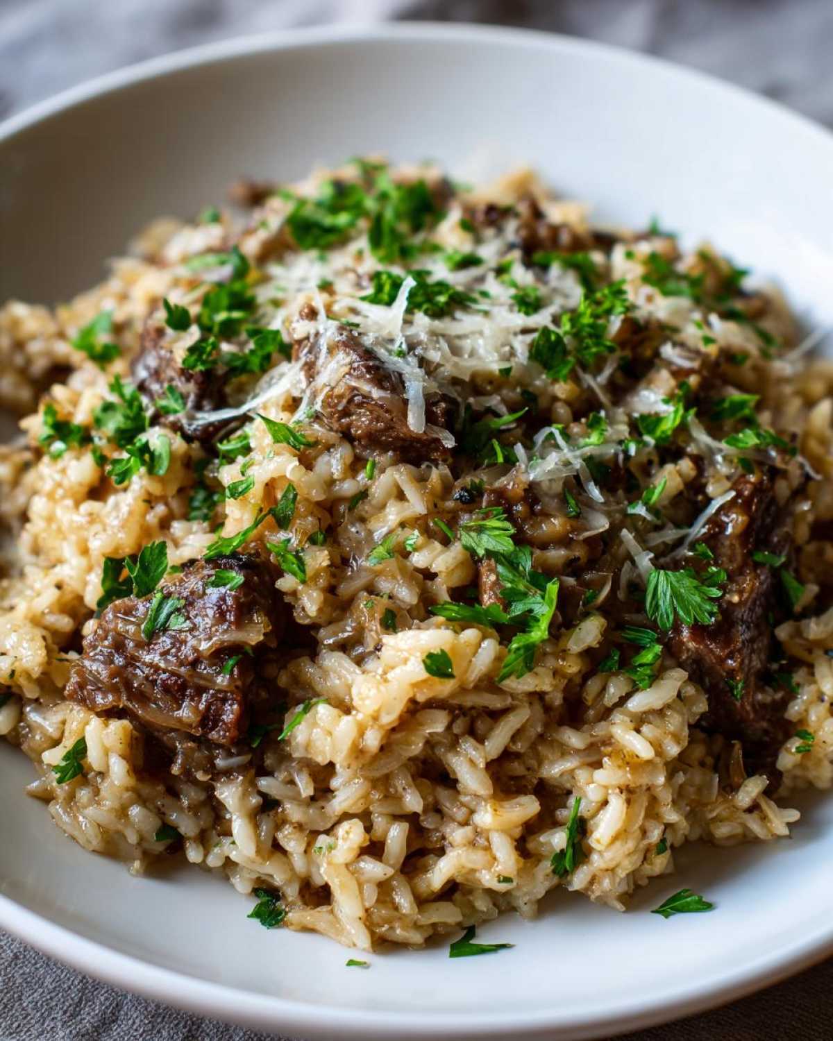 Close-up of creamy french onion butter rice topped with braised beef chunks, grated parmesan, and fresh parsley.