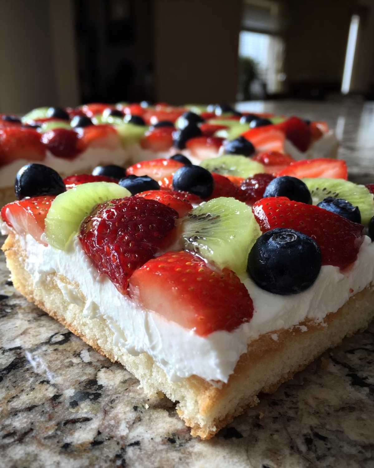 Close-up of a slice of fruit pizza sugar cookie topped with whipped cream, strawberries, blueberries, and kiwi.