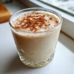 A glass of creamy, frothy coquito topped generously with ground cinnamon, sitting on a white surface.