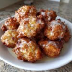 A pile of freshly fried, golden brown apple fritter bites drizzled with a sweet white glaze.