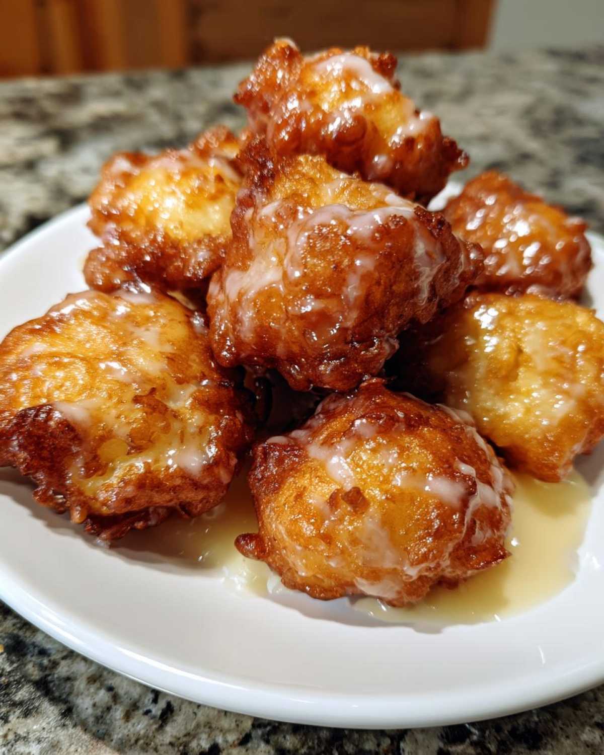 A stack of golden brown, freshly fried apple fritter pieces drizzled with a sweet white glaze, resting on a white plate.