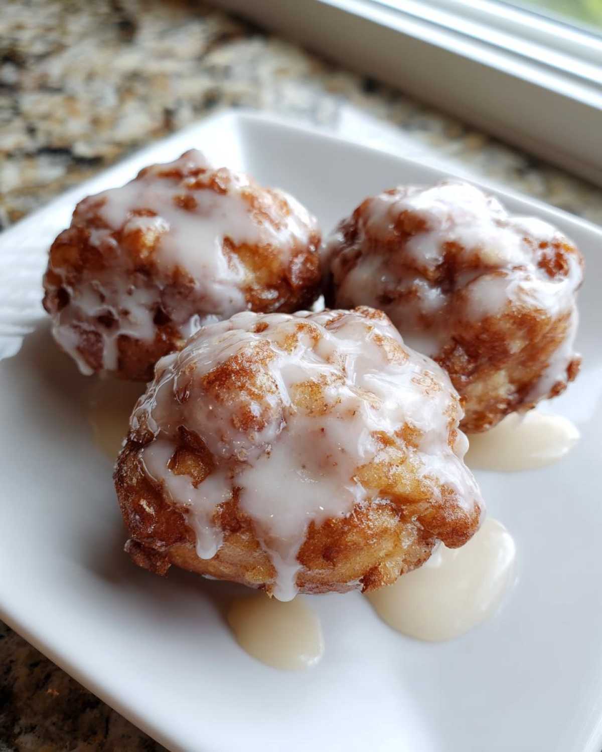 Three golden brown baked apple fritters drizzled generously with white glaze, sitting on a white plate.