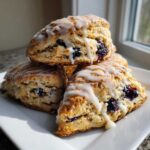 A stack of three freshly baked blueberry scones drizzled with a thick white glaze, sitting on a white plate.