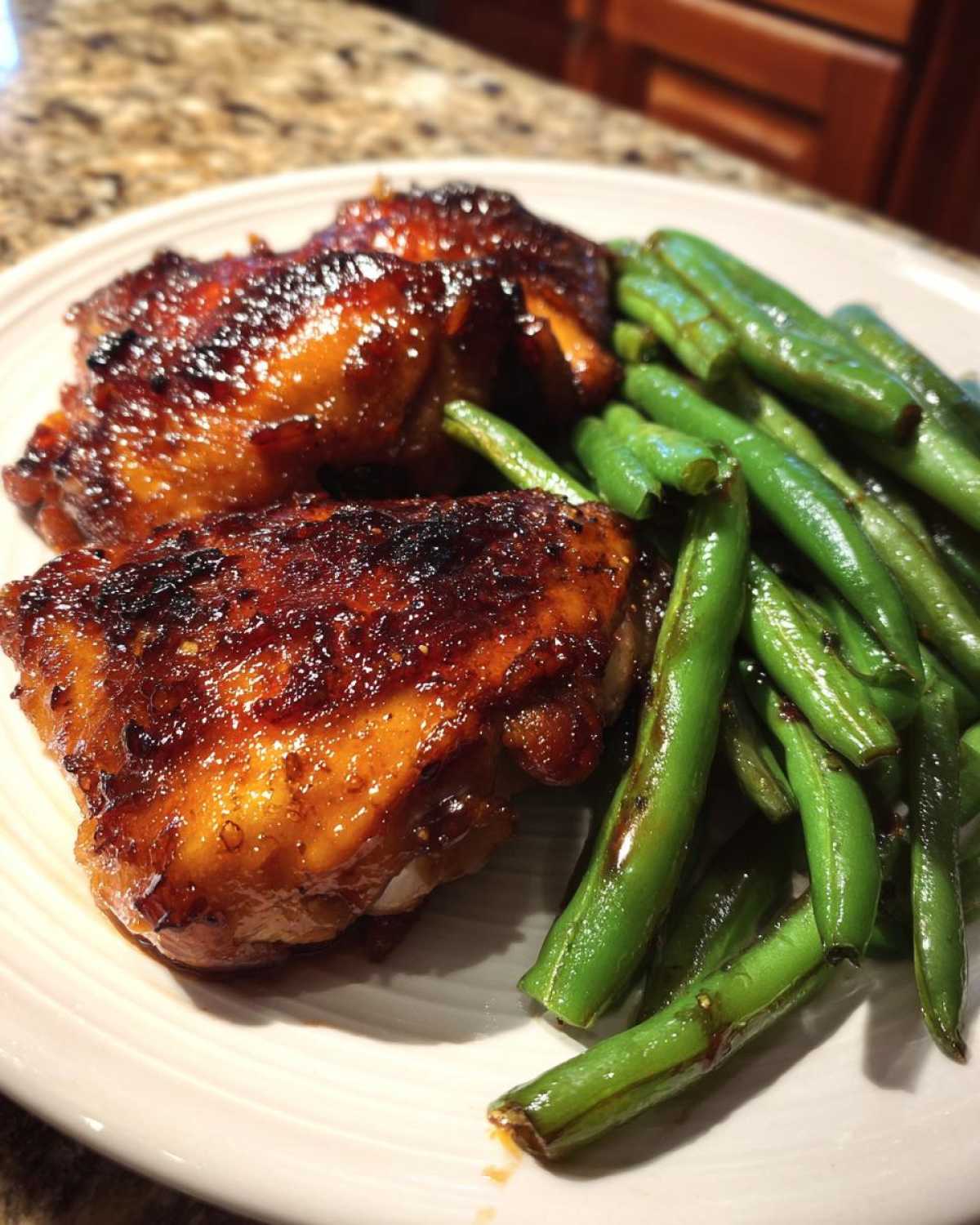Two pieces of glazed, caramelized chicken served next to bright green beans on a white plate.