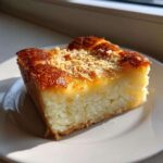 A square slice of moist cassava cake with a rich, golden-brown, slightly caramelized top sprinkled with crumbs.
