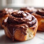 Close-up of a freshly baked chocolate cinnamon roll topped with thick, dripping chocolate frosting.