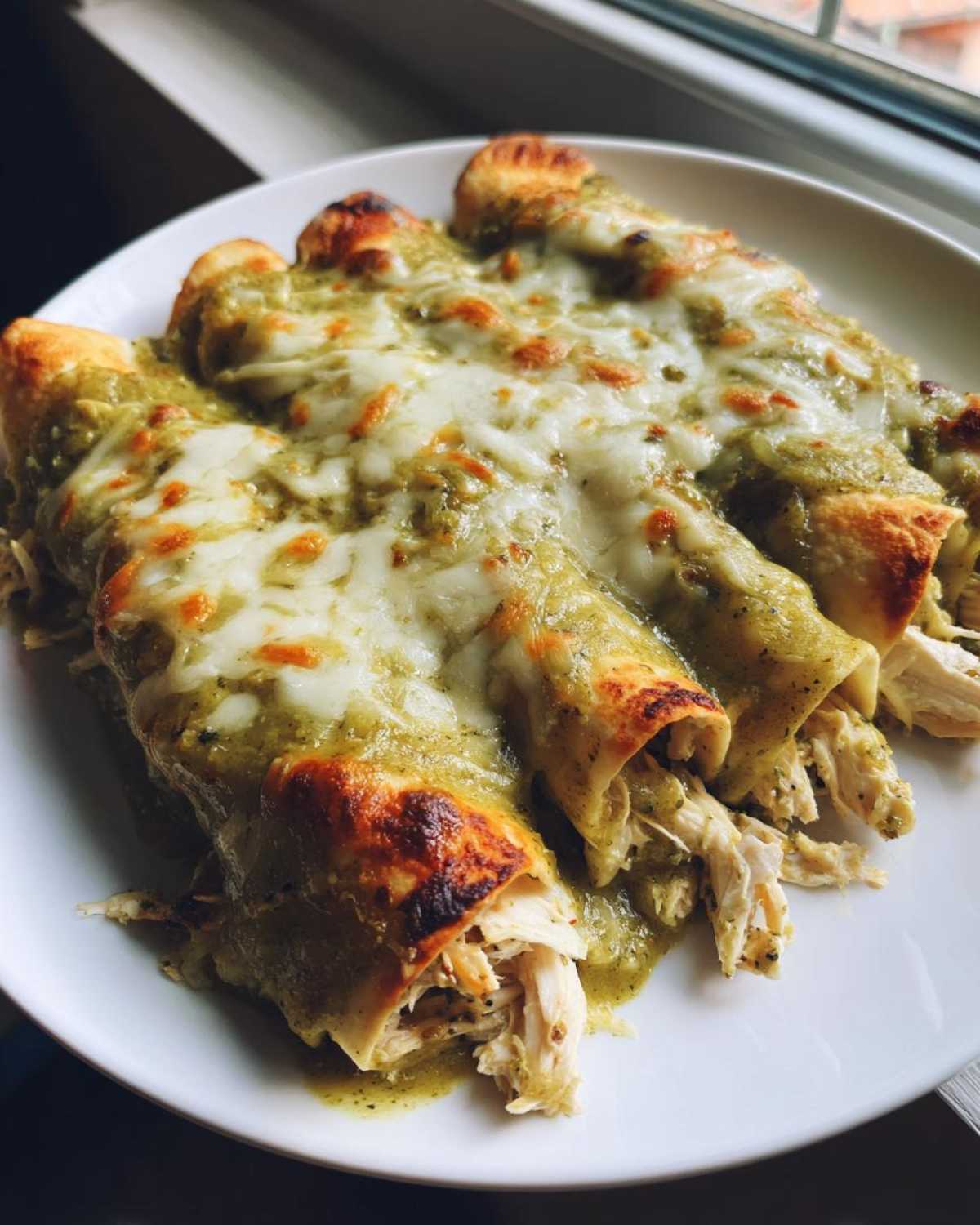 Three baked green chile chicken enchiladas smothered in green sauce and melted, slightly browned cheese on a white plate.