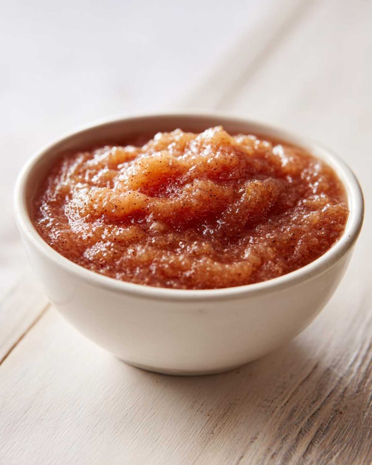 Close-up of chunky, spiced applesauce served in a small white bowl on a light wooden surface.