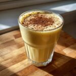 A close-up of homemade eggnog in a clear glass, topped with a dusting of cinnamon.