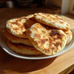 A stack of freshly cooked, golden-brown pita bread with charred spots resting on a white plate.