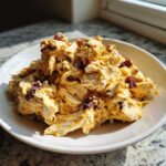 A mound of creamy honey mustard chicken salad mixed with shredded chicken and topped with dark bacon pieces, served on a white plate.