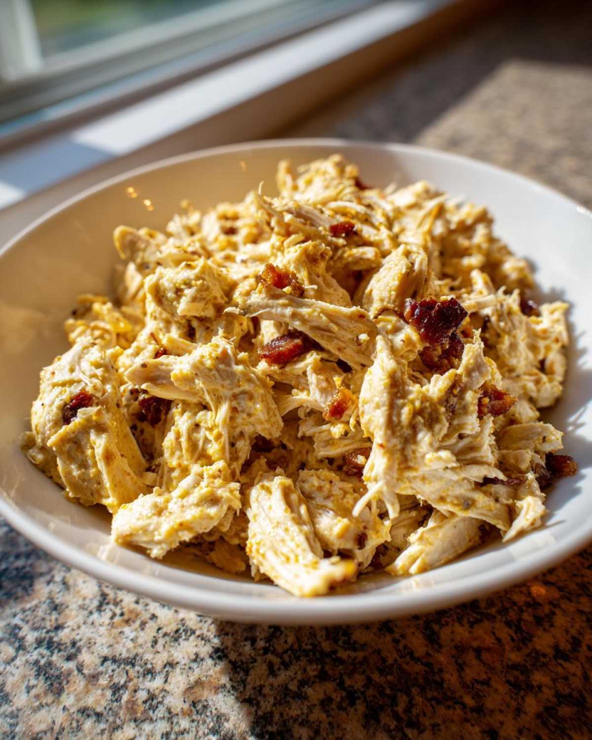Close-up of shredded chicken mixed with a creamy dressing in a white bowl, resembling honey mustard chicken salad with bacon.