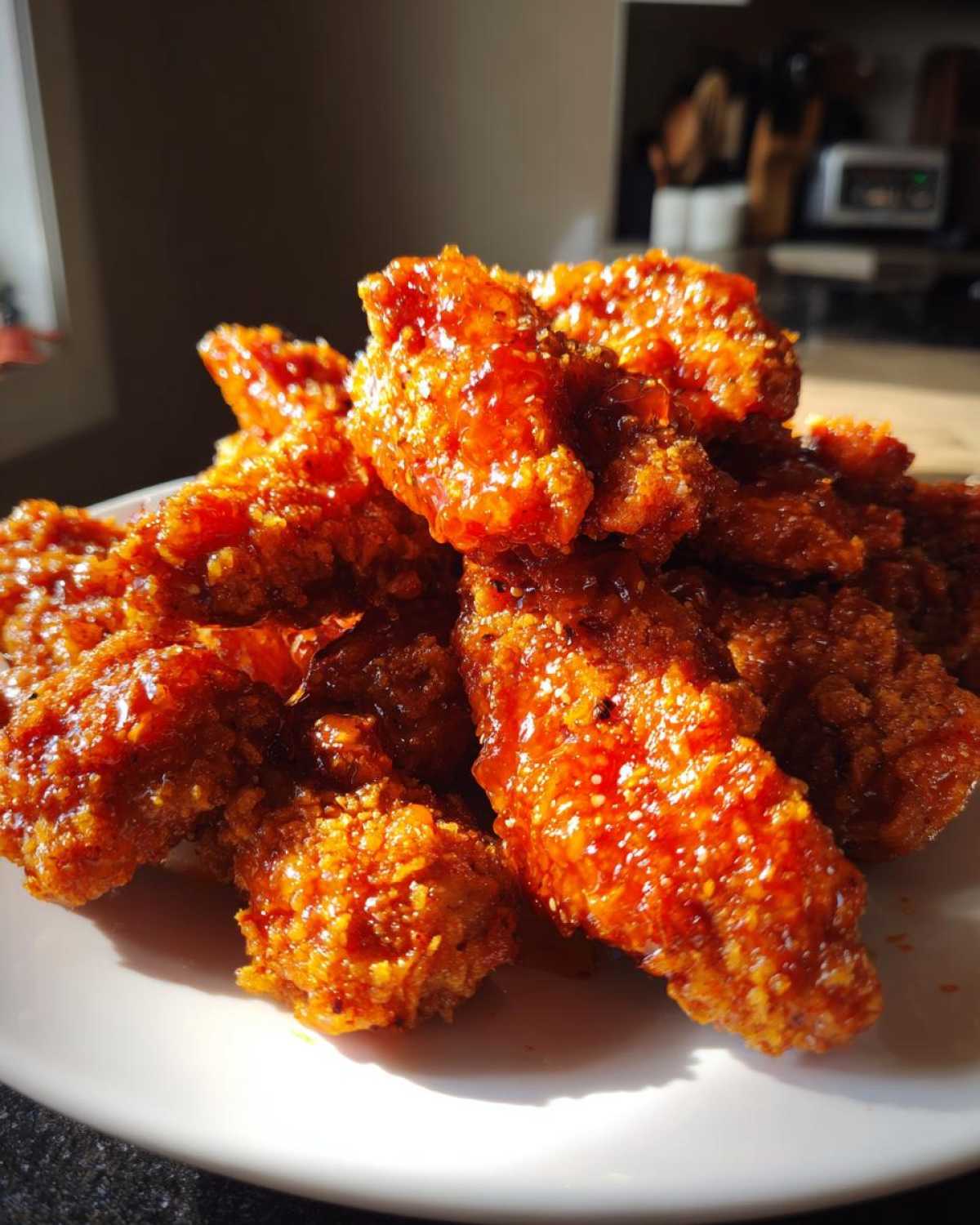 A close-up of crispy, glazed hot honey chicken tenders piled on a white plate, glistening under natural light.