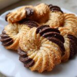 Close-up of freshly baked Italian butter cookies, piped into swirls and partially dipped in dark chocolate, sprinkled with coconut.