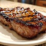 Close-up of a thick, caramelized, and juicy pork steak recipe piece resting on a white plate.