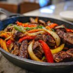Close-up of sizzling steak fajitas with grilled steak strips, onions, and colorful bell peppers in a cast iron skillet.
