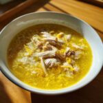 A close-up of a white bowl filled with vibrant yellow Lemon Ginger Turmeric Chicken and Rice Soup, featuring shredded chicken and rice.