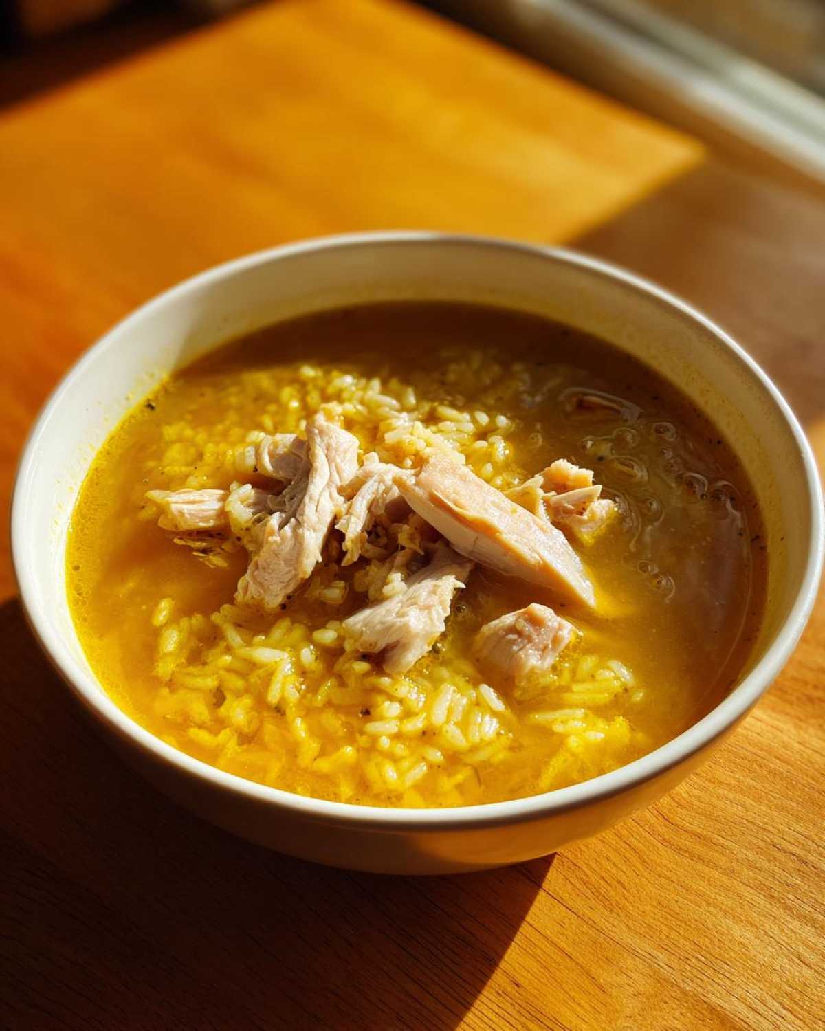 A close-up of a bowl of vibrant yellow Lemon Ginger Turmeric Chicken and Rice Soup topped with shredded chicken.