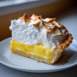 A close-up of a generous slice of lemon meringue pie showing bright yellow filling and tall, toasted meringue peaks.