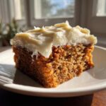A close-up of a square slice of moist applesauce cake topped with thick, creamy white frosting.
