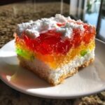 A colorful slice of jello poke cake featuring layers of bright red, orange, and green gelatin over a white cream layer and cake base.