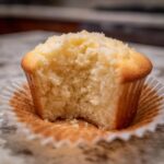 Close-up of a vanilla cupcakes with a bite taken out, showing the moist, fluffy interior and crumb topping.